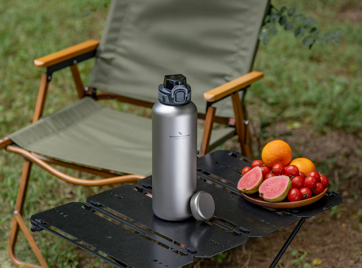 Boundless Voyage titanium water bottle and dual lids displayed on camp table beside fresh fruit