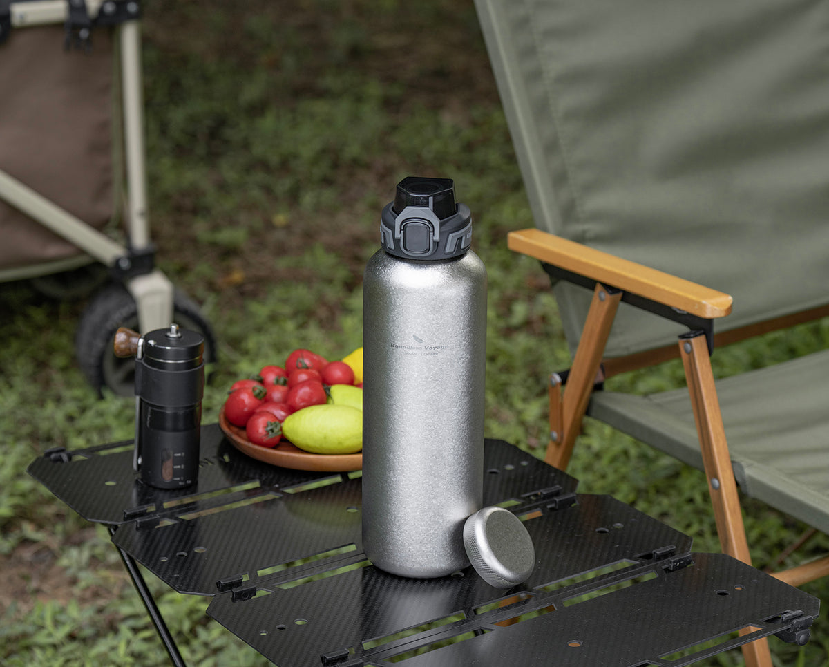 Crystallized silver titanium bottle and dual lids on camp table next to manual coffee grinder